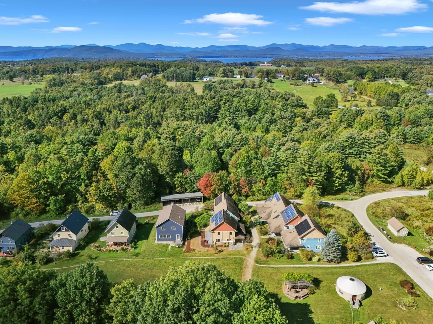 CVC from above: colorful farmhouse-style homes with solar panels around a green, the yurt and garden in front, woods and fields beyond, and Lake Champlain and the Adirondacks on the horizon